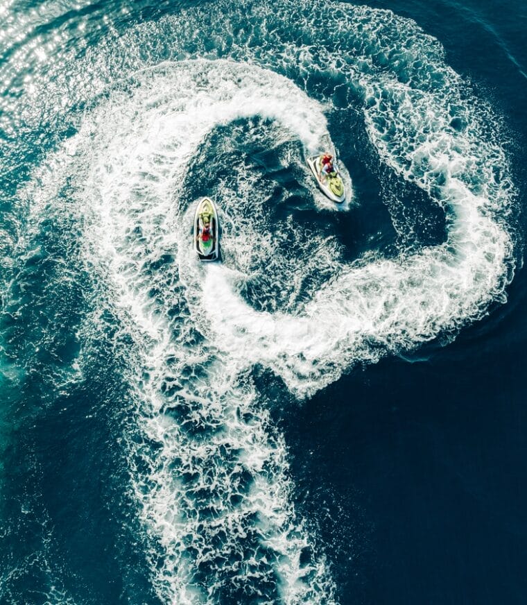 Aerial view of two people on two jet skis, creating a large circular wave pattern in the ocean.