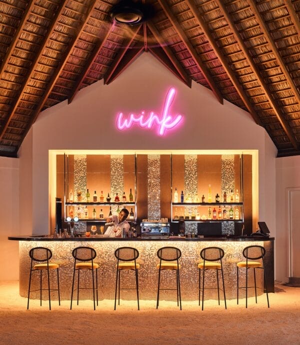 Stylish tropical bar with a thatched roof and a neon 'Wink' sign, featuring a sleek counter with mosaic tiling, a bartender preparing drinks, and high stools lined up in front, illuminated by warm lighting.