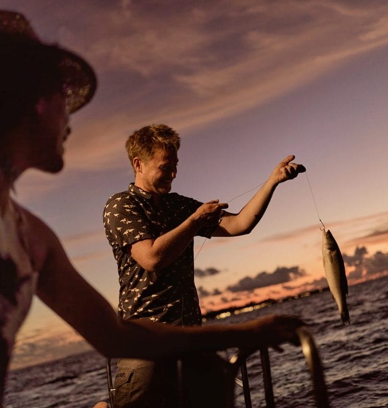 Man holding a freshly caught fish while fishing at sunset, with a woman watching nearby. The sky is painted in warm hues of orange and purple, creating a peaceful and scenic backdrop over the ocean.