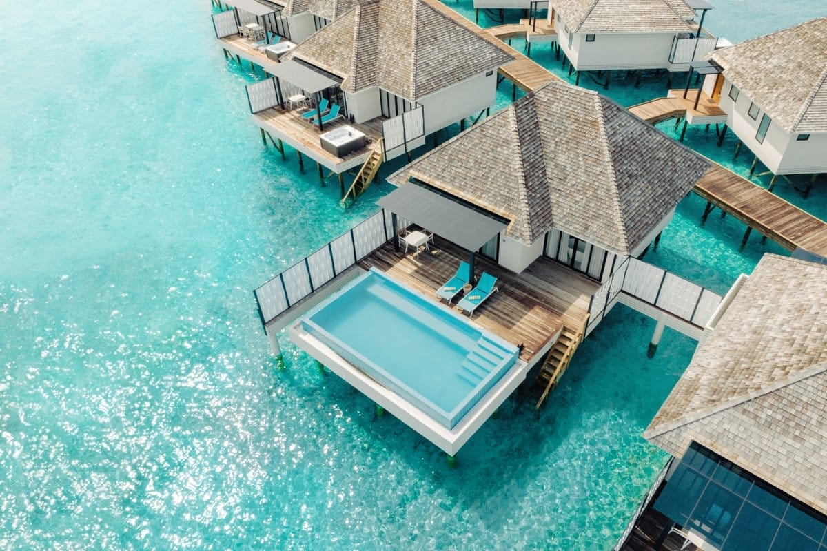 Aerial view of a luxurious overwater villa with a private infinity pool and wooden deck, situated above clear turquoise waters in a tropical lagoon.