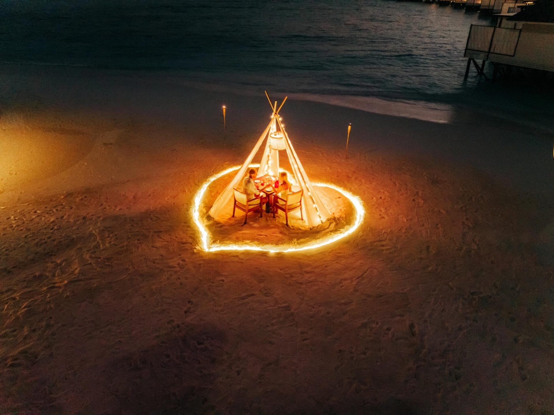 Heart-shaped dining setup on the beach, glowing under the night sky, perfect for a romantic evening.