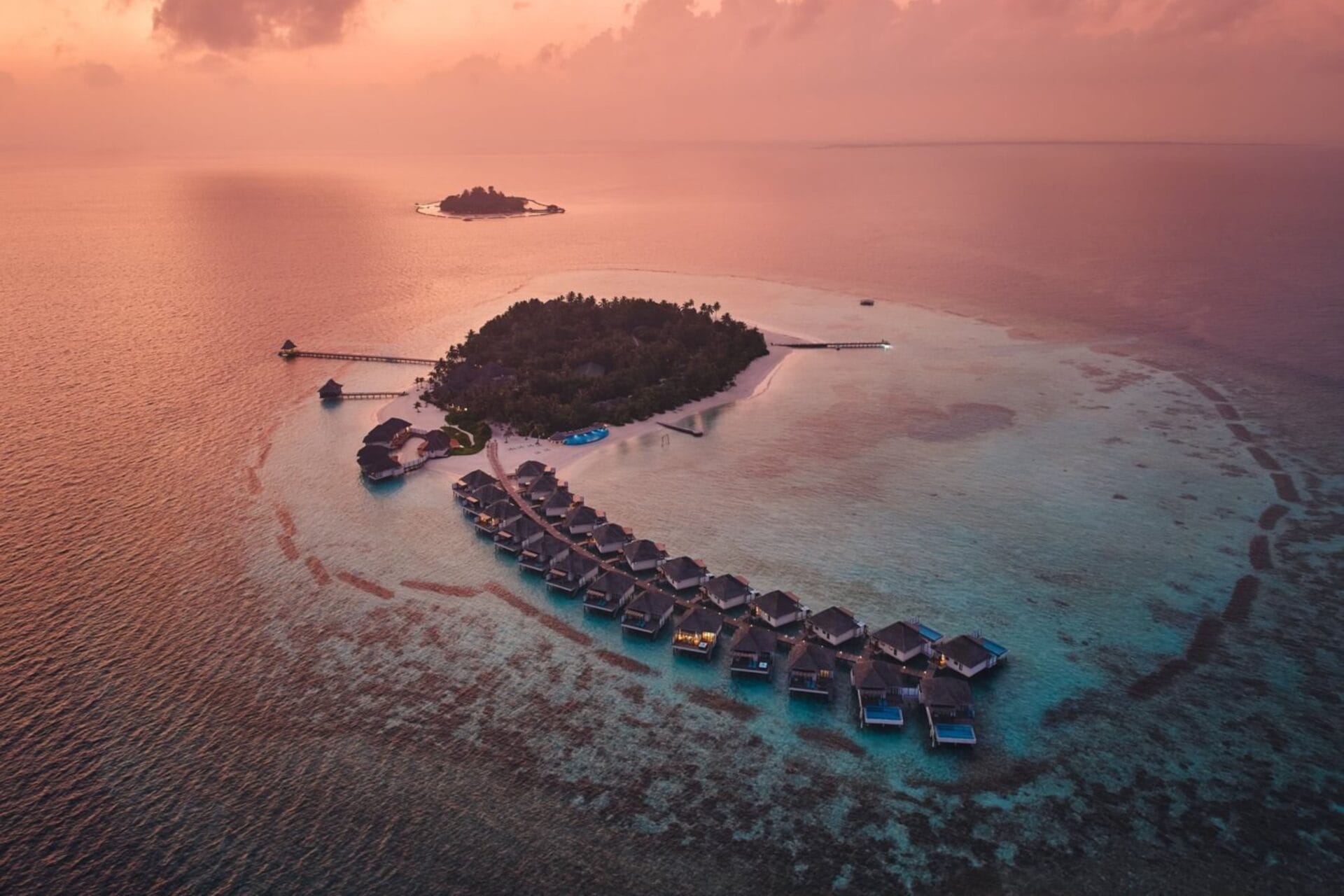 Aerial view of overwater villas at sunset with vibrant colors reflecting on the ocean.