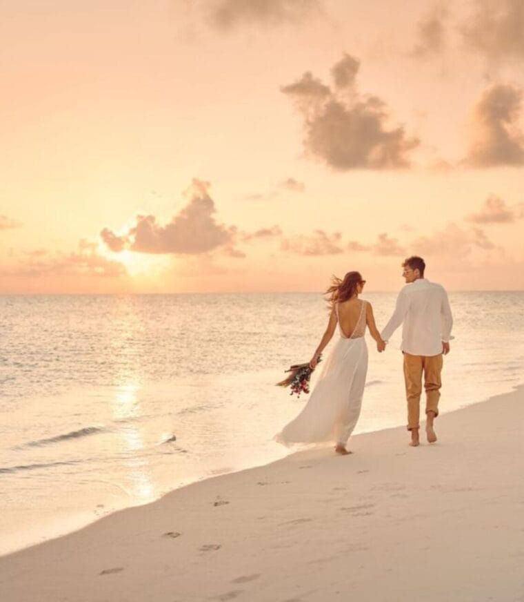 A couple holding hands while walking barefoot along a sandy beach at sunset. The woman is wearing a flowing white dress and carrying a small bouquet, while the man is dressed casually in a white shirt and tan pants. The sky is bathed in soft pink and orange hues, creating a romantic atmosphere.