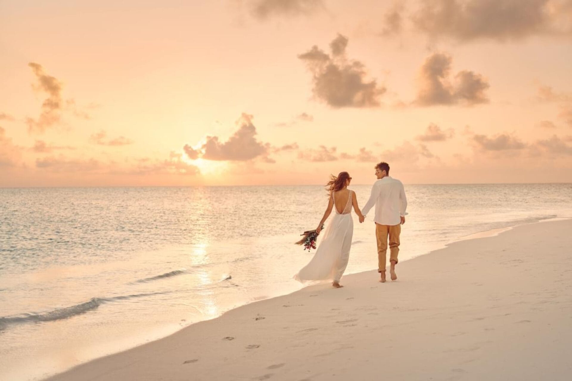 A couple holding hands while walking barefoot along a sandy beach at sunset. The woman is wearing a flowing white dress and carrying a small bouquet, while the man is dressed casually in a white shirt and tan pants. The sky is bathed in soft pink and orange hues, creating a romantic atmosphere.