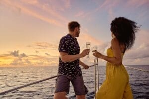 A couple standing on the deck of a boat during a sunset cruise, each holding a glass of champagne. The man is wearing a patterned shirt and shorts, while the woman is in a yellow dress. The sky is painted in shades of pink and orange, reflecting the peaceful ocean below