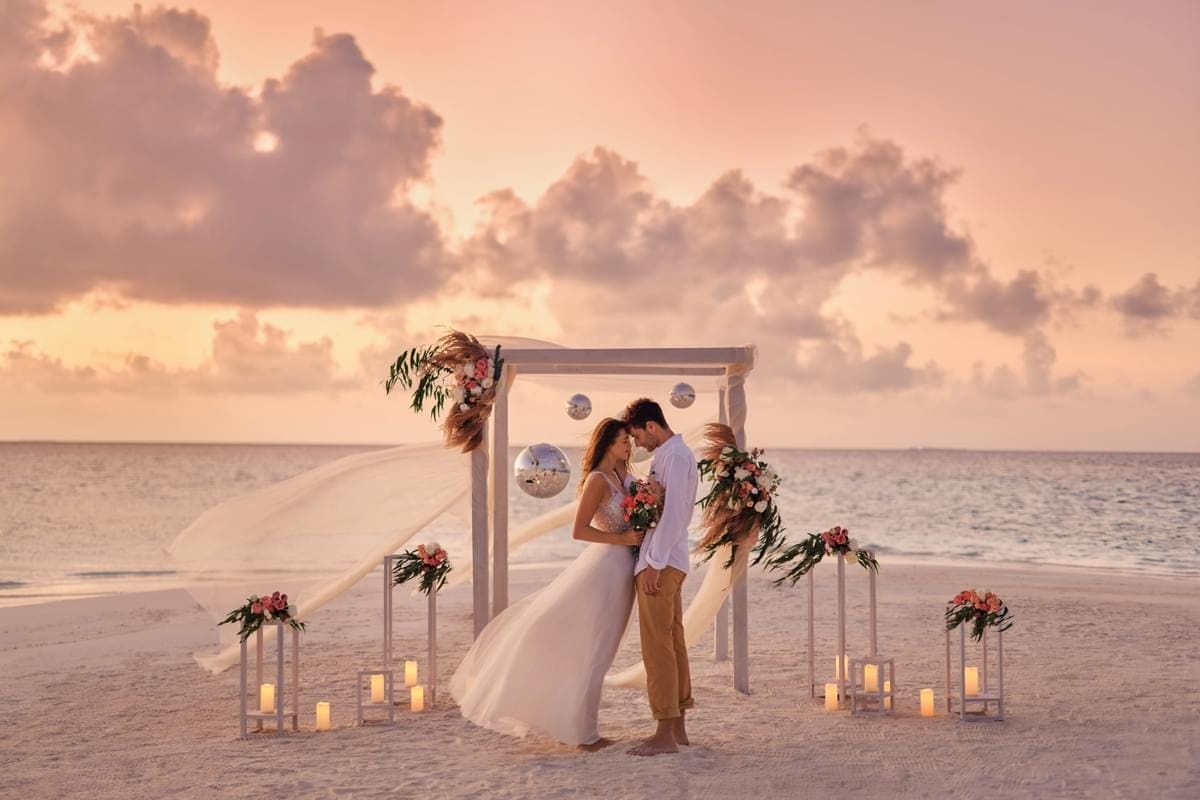 A couple standing under a decorated wedding arch on a sandy beach at sunset. The bride is wearing a white dress and holding a bouquet, while the groom is in a white shirt and tan pants. The arch is adorned with flowers, fabric, and lanterns, creating a dreamy and intimate setting.