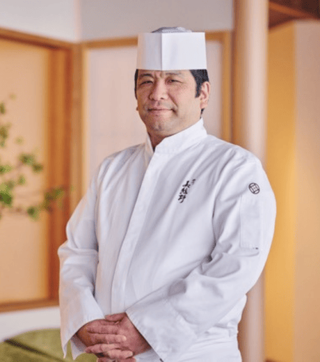 A chef in a white uniform smiling in a professional kitchen environment.