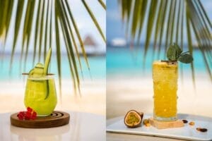 Two tropical drinks set against a backdrop of palm fronds and turquoise ocean. On the left, a bright green cucumber-based cocktail garnished with a cucumber ribbon and red berries. On the right, a golden passionfruit mocktail with a sprig of mint, accompanied by a halved passionfruit and tropical garnishes.